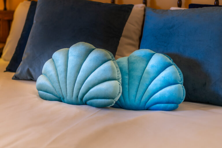 Cojines en forma de concha, color turquesa, sobre una cama con almohadas azules y beige, creando un ambiente acogedor.