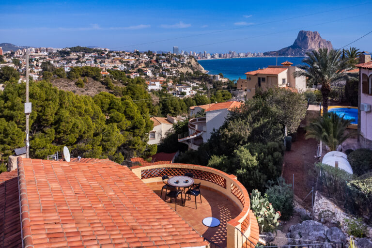 Vista panorámica de una zona montañosa con casas, árboles, y un gran peñón junto al mar bajo un cielo despejado.