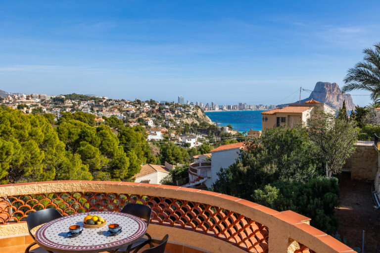 Vista panorámica de casas sobre colinas verdes, mar azul, una montaña al fondo, y mesa con café en un balcón semicircular.