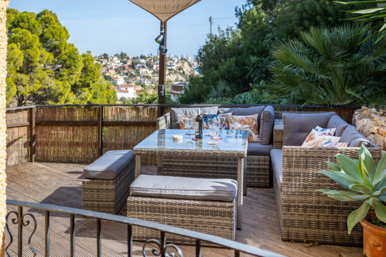 Terraza con muebles de mimbre, cojines estampados, mesa de vidrio, botella de vino, rodeada de plantas y vista a ciudad colina.