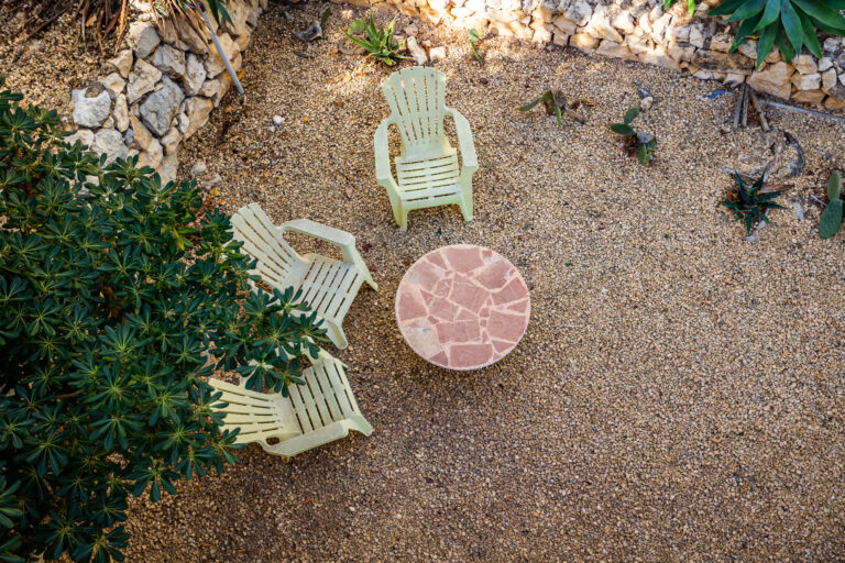 Vista desde arriba de un patio con tres sillas de plástico verdes, mesa redonda de mosaico y plantas alrededor. Suelo de grava.