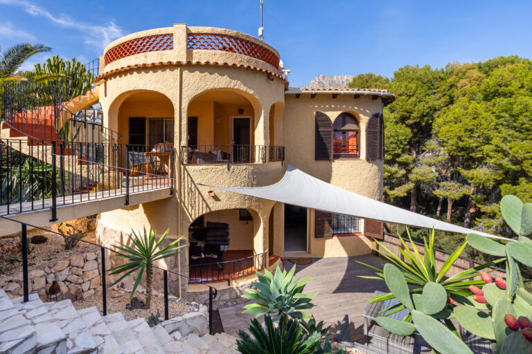 Casa estilo mediterráneo con arcos, terraza elevada, ventanas con persianas, plantas tropicales y sendero de piedra. Cielo azul.