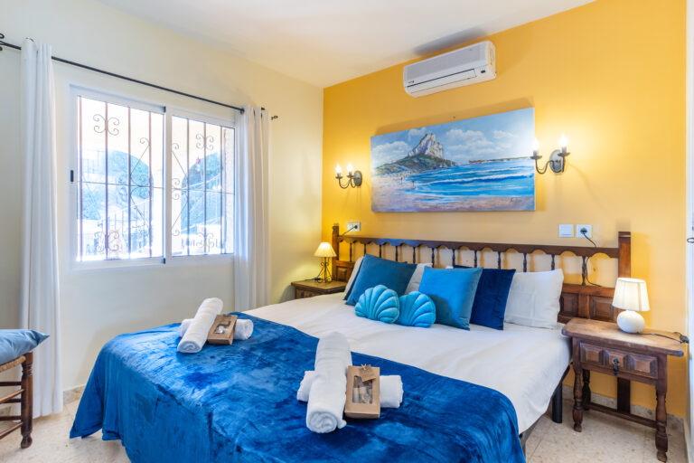 Habitación luminosa con cama doble, colcha azul, cojines turquesa. Ventana con cortinas blancas, pared amarilla y pintura de playa.