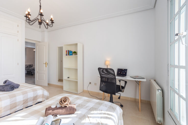 Habitación luminosa con dos camas individuales, escritorio con silla y lámpara, estantería blanca y ventana amplia con luz natural.