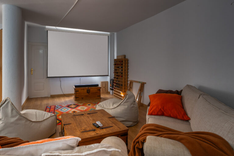 Sala de estar con sofá beige, cojines naranjas, alfombra colorida, pantalla de proyección y mueble de madera.