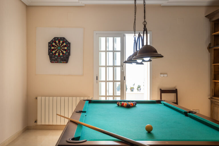 Sala de juegos con mesa de billar verde, lámparas colgantes y diana de dardos en la pared junto a una puerta corrediza.