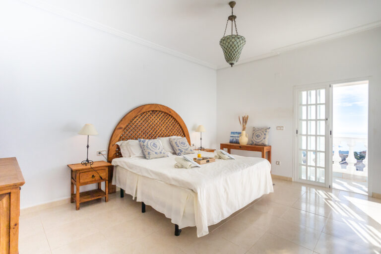 Habitación luminosa con cama doble, cabecero de madera, mesitas, lámparas y terraza con vista al mar; tonos blancos y azules.