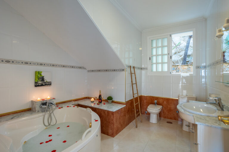 Baño con bañera de hidromasaje, pétalos rojos flotantes, velas, ventana con árboles, lavabo y bidé, ambiente relajante.