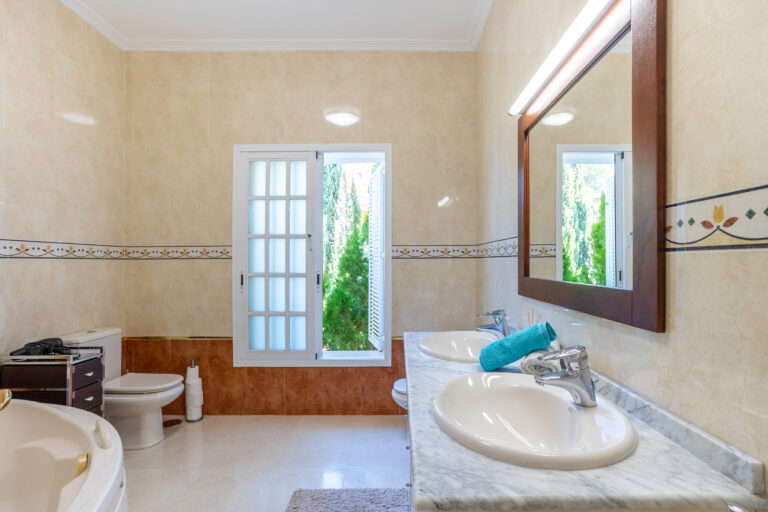 Baño luminoso con dos lavabos, espejo grande, bañera redonda, ventana amplia con vista a árboles y paredes decoradas.