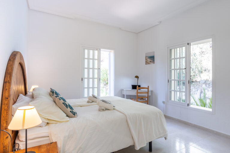 Dormitorio luminoso con cama doble de cabecera de madera, dos ventanales grandes, escritorio blanco y lámpara de mesa.