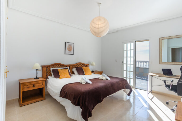 Habitación luminosa con cama doble, cabecero de madera, lámparas en mesitas, escritorio con silla y puerta a balcón.