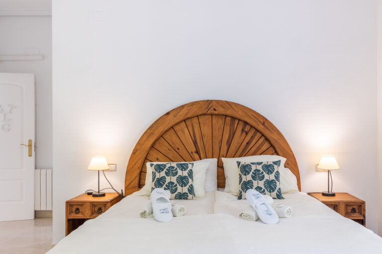 Cama con cabecera de madera semicircular, almohadas con fundas de hojas verdes, lámparas y mesitas de noche de madera.