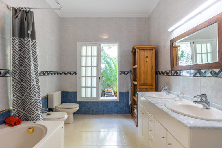 Baño amplio con ducha y bañera, lavamanos doble, azulejos azules y blancos. Armario de madera y ventana a jardín externo.