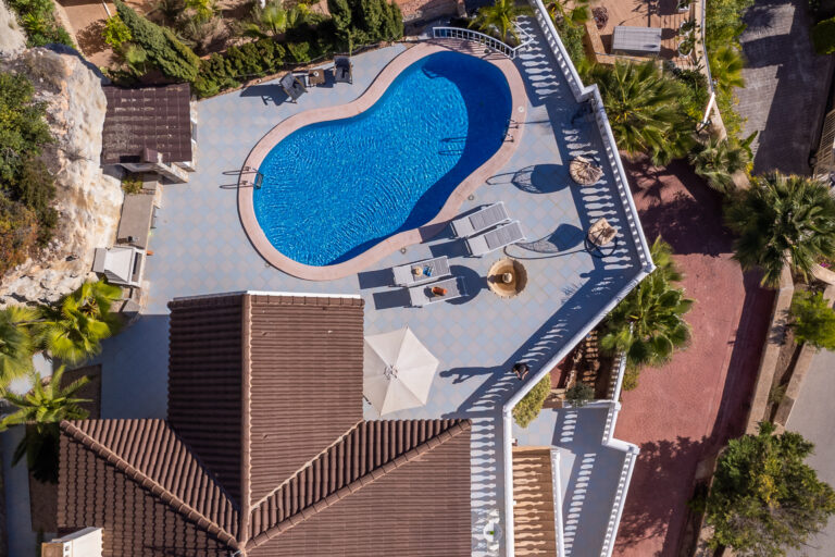 Vista aérea de una piscina con forma irregular rodeada de tumbonas grises, sombrillas y un tejado marrón. Plantas verdes alrededor.