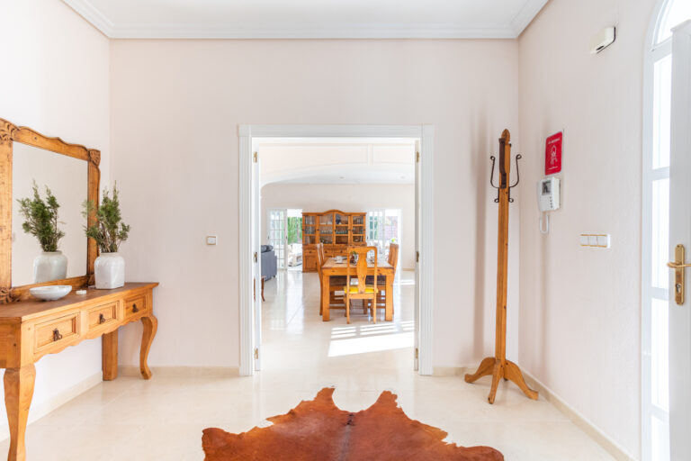 Amplio recibidor con espejo y consola de madera, alfombra de piel, percha y vista a un comedor iluminado.