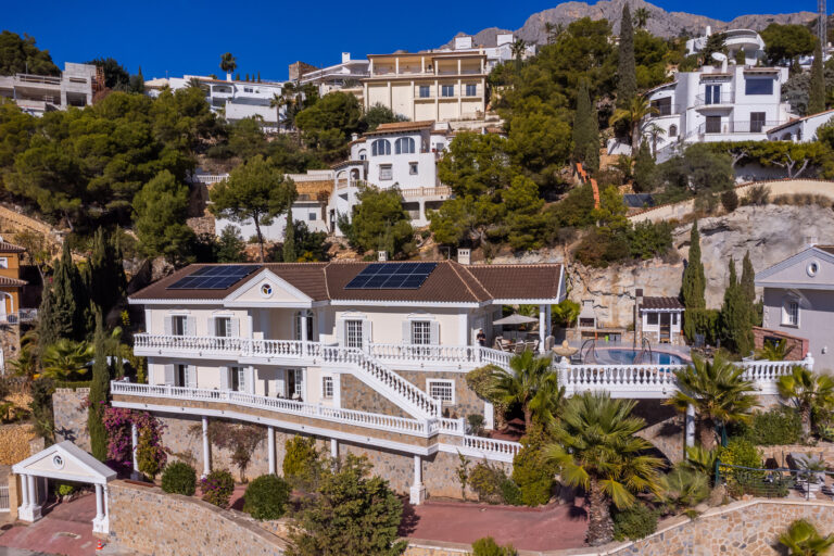 Casa blanca con paneles solares, rodeada de vegetación y otras viviendas en ladera de colina, cielo azul claro.