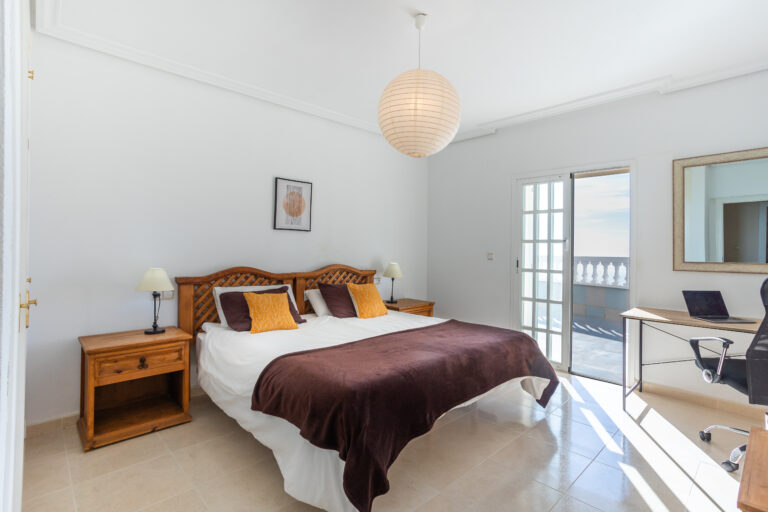 Dormitorio luminoso con cama doble, cabecera de madera, lámparas de noche, escritorio con silla, y puerta a un balcón exterior.