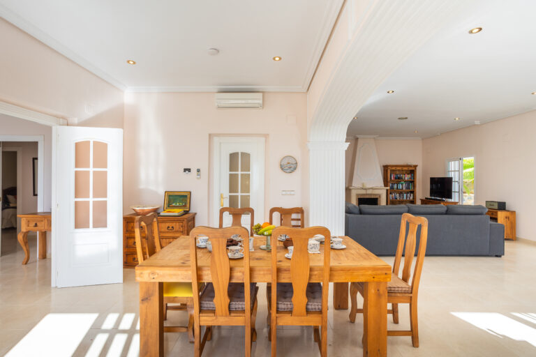 Sala espaciosa con muebles de madera clara, mesa de comedor, sofás grises, chimenea, estantería y ventanas amplias. Ambiente luminoso.