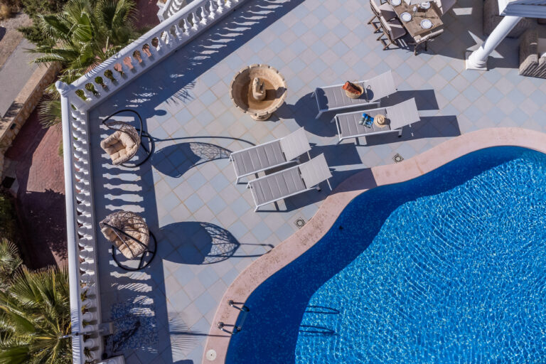 Vista aérea de piscina azul, con tumbonas blancas, sillas colgantes y una mesa con vajilla en un patio soleado.