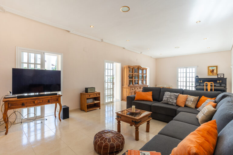 Amplio salón con sofá gris y cojines naranjas, televisor sobre mesa de madera y piano negro en el fondo.