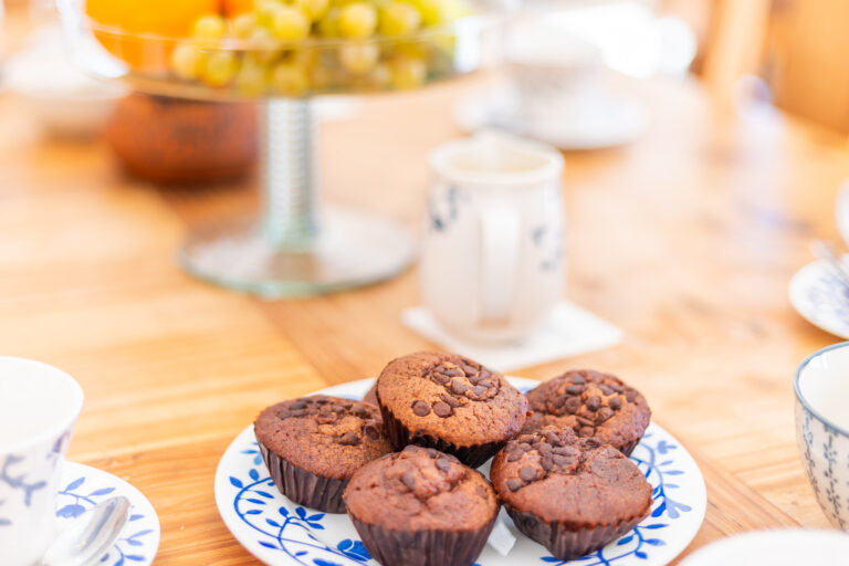 Muffins de chocolate en un plato decorativo azul sobre mesa de madera, jarra y frutas en el fondo.
