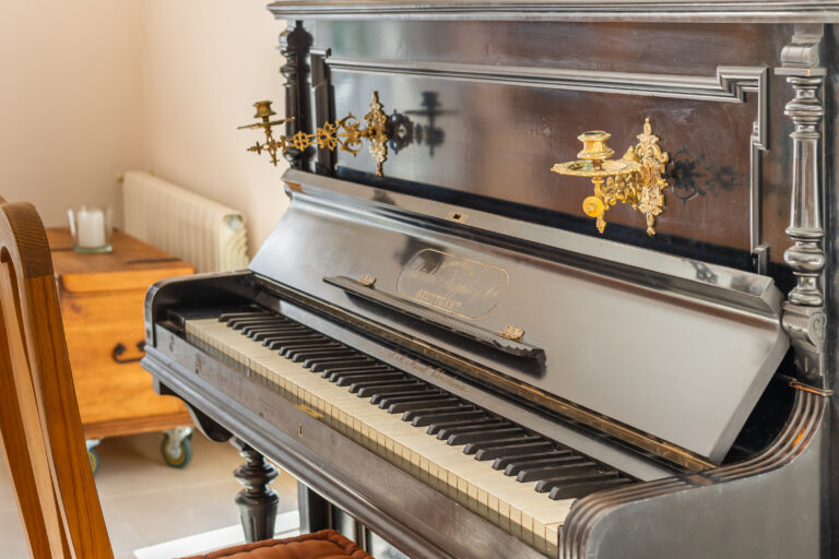 Antiguo piano vertical negro con candelabros dorados decorativos, teclas blancas y negras, situado junto a una silla de madera.
