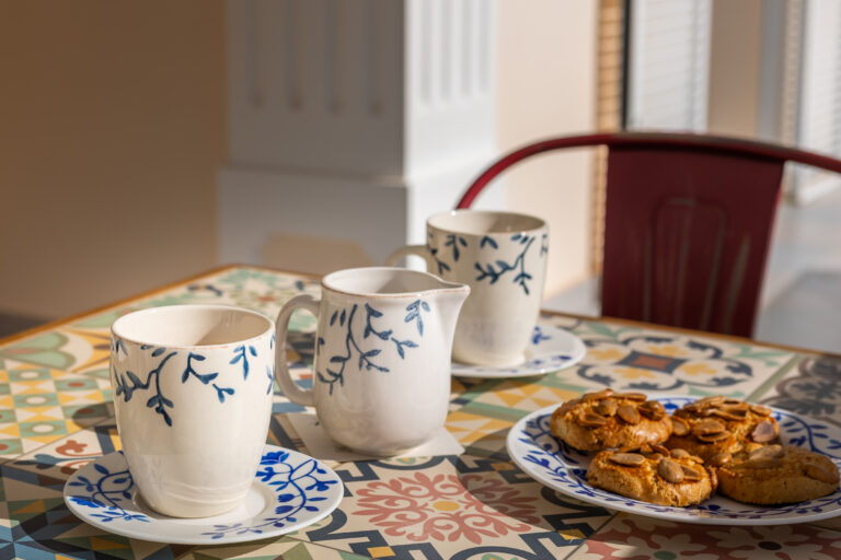 Tazas y jarra de cerámica blanca con diseño azul sobre mesa colorida, plato con galletas cubiertas de almendras.