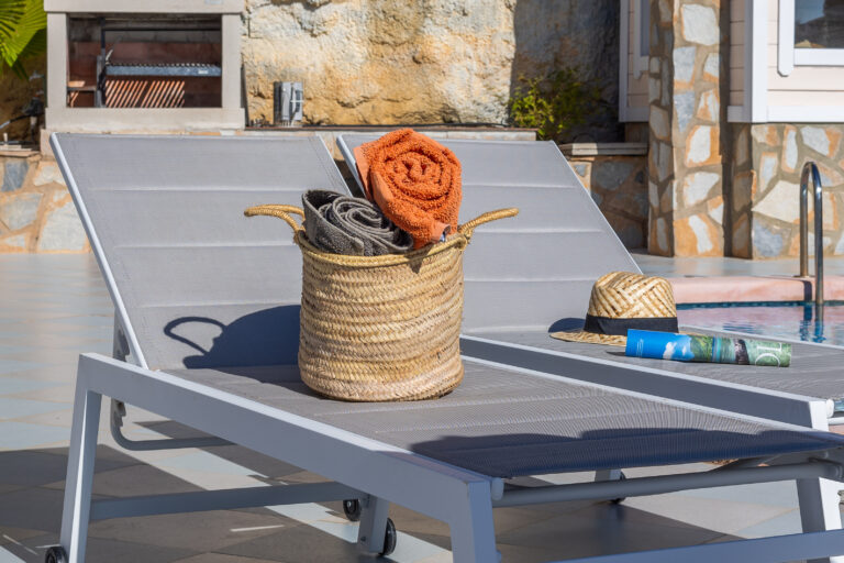 Dos tumbonas grises junto a piscina. Canasta con toallas, sombrero de paja, revista. Asador de ladrillo al fondo.