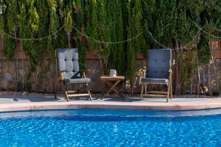Dos sillas grises junto a una mesa pequeña frente a una piscina azul. Al fondo, setos verdes y cadena de luces.