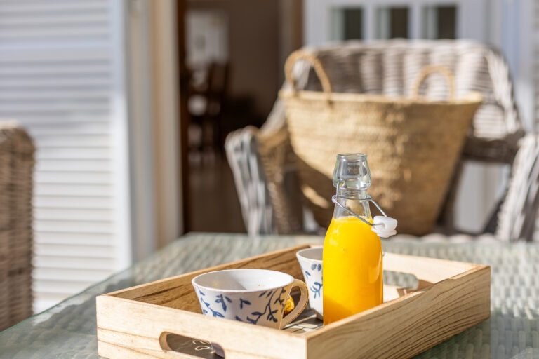 Botella de jugo naranja y tazas blancas con motivos azules en bandeja de madera sobre mesa de vidrio. Ambiente soleado, sillas de mimbre.