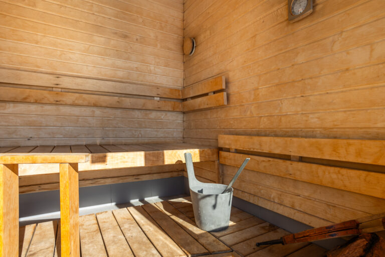 Interior de sauna de madera clara, con bancos escalonados, un cubo de metal y una pala. Luz solar entrando. Termómetro en la pared.