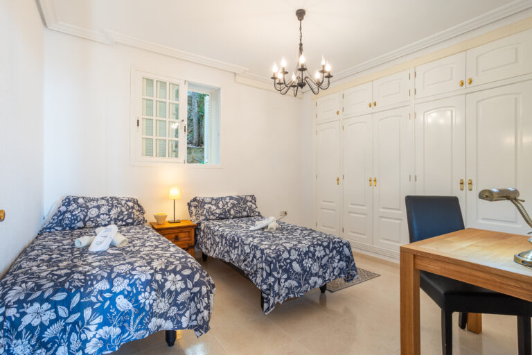 Habitación con dos camas individuales, cobertores florales azules, ventana, lámpara en mesita, armarios blancos y escritorio.