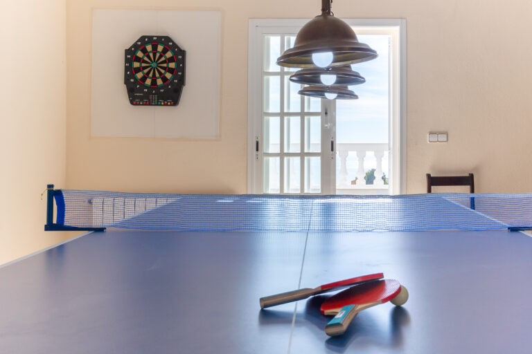 Mesa de ping pong con paletas y pelotas sobre ella, diana en la pared. Puerta de vidrio con vista exterior y lámpara colgante.