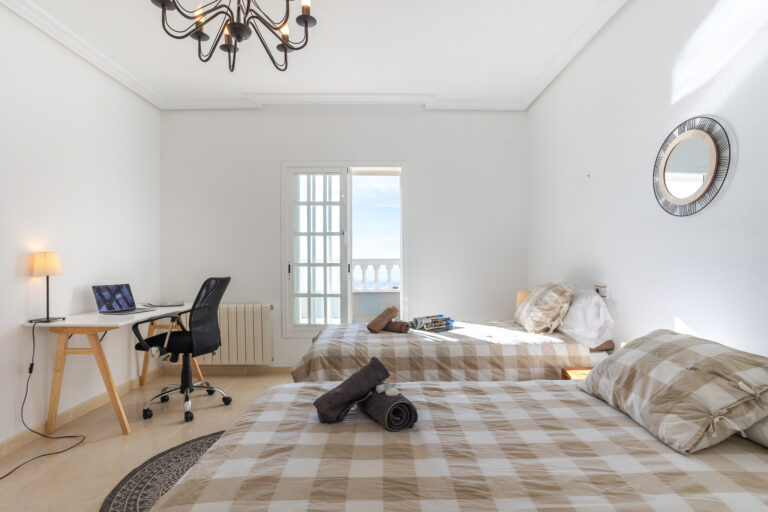 Habitación luminosa con dos camas de cuadros beige, escritorio con lámpara y silla negra, ventana con vistas y candelabro negro.